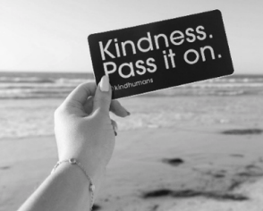 Hand hält Schild mit der Aufschrift „Kindness. Pass it on.“ vor einem Strand.